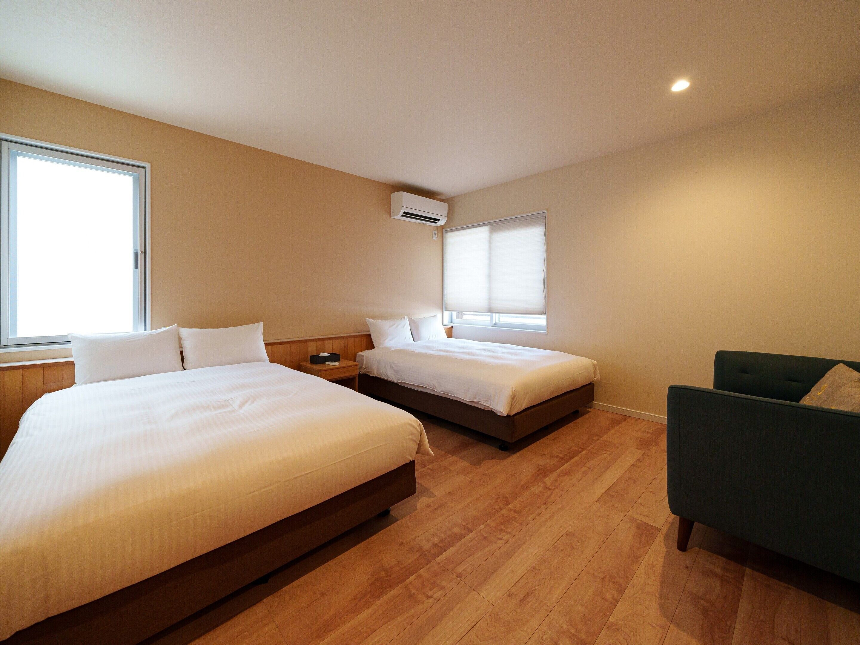 A hotel room with two beds, a green armchair, wooden flooring, and a window with blinds.