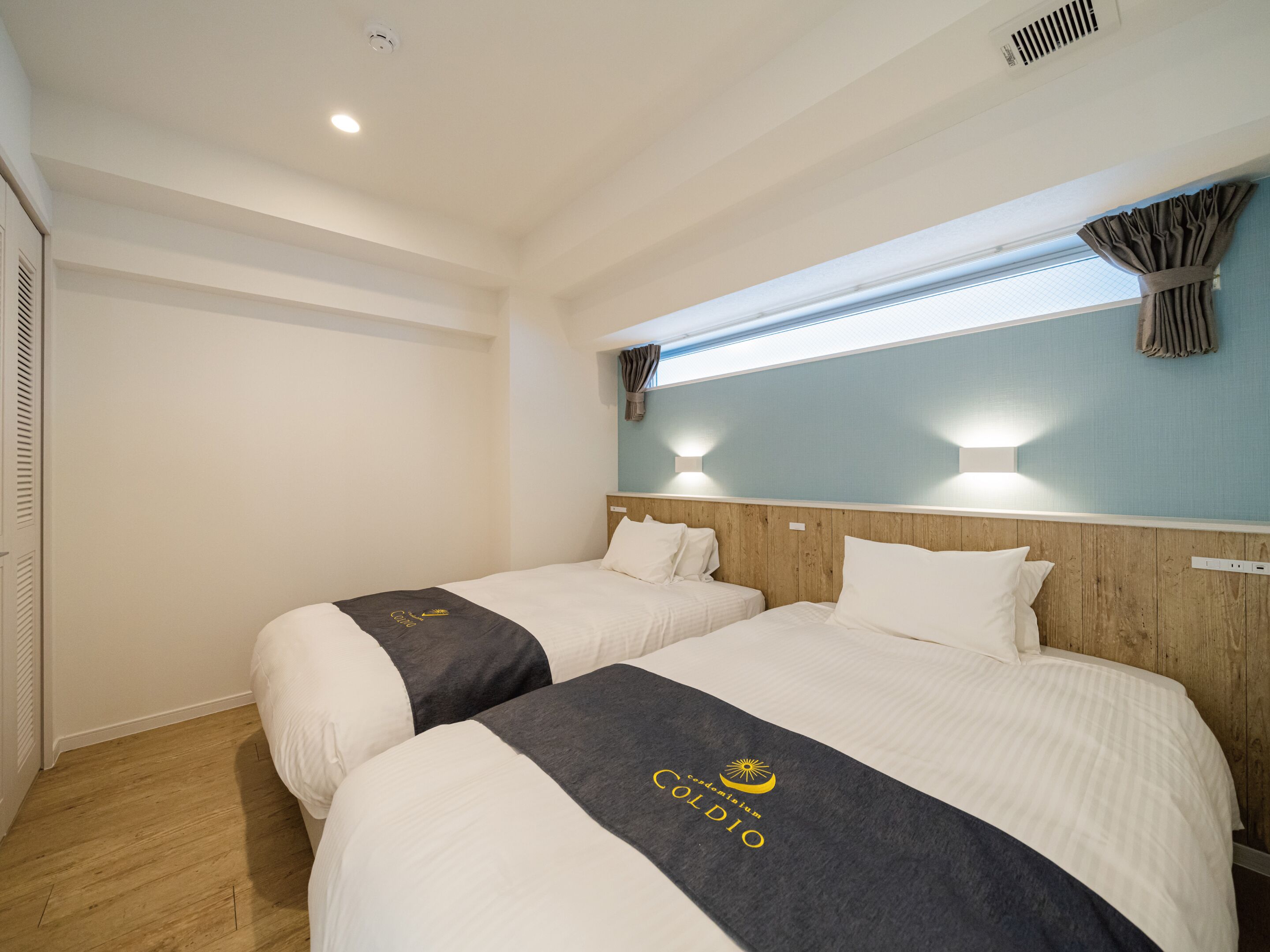 A hotel room with two beds, a wooden headboard, a window with curtains, and a ceiling vent.