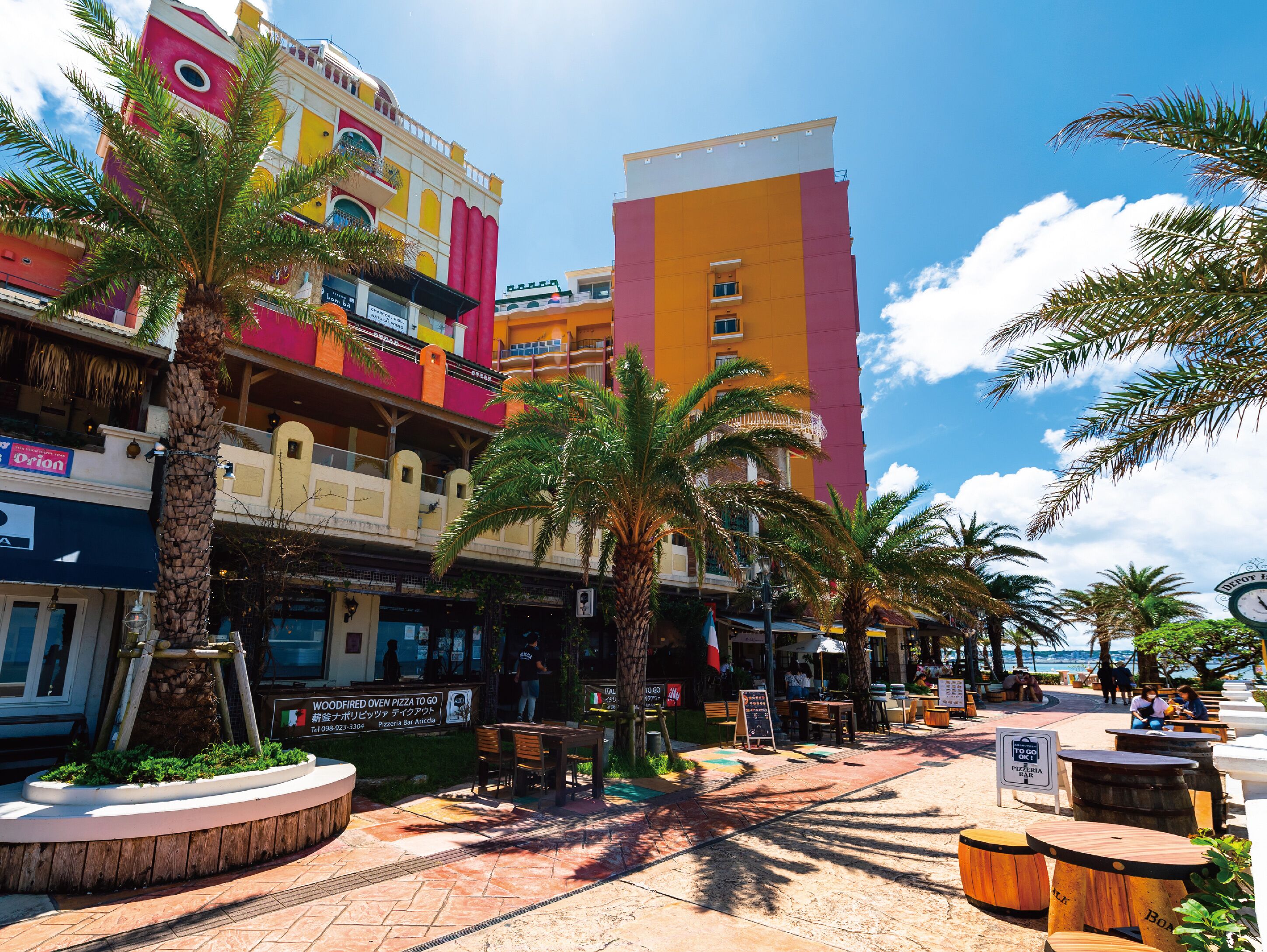 A vibrant outdoor plaza with palm trees, wooden tables, and a variety of shops and restaurants.