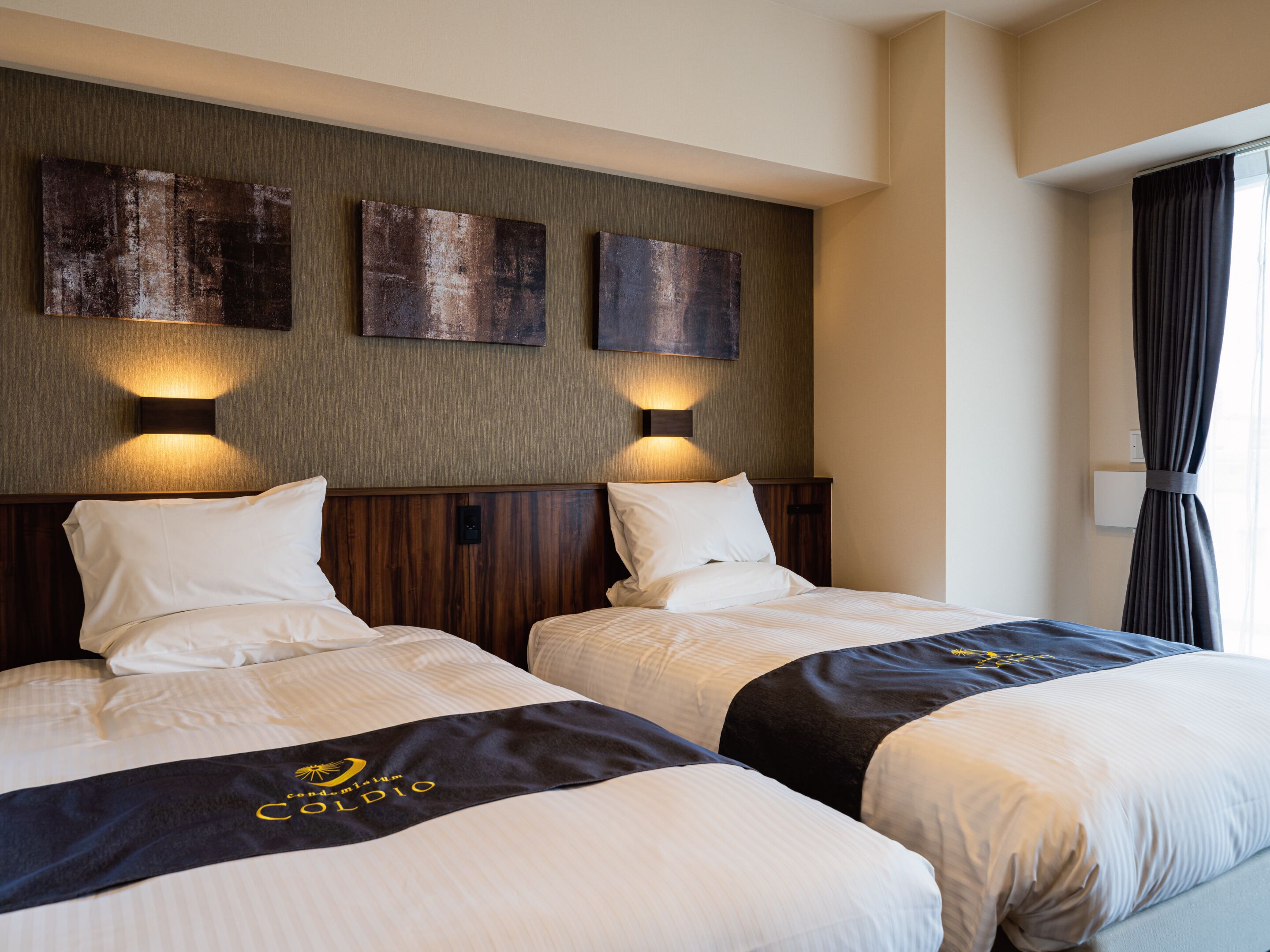 A hotel room with two beds, each with a headboard and pillows. There are wall-mounted light fixtures above the beds.