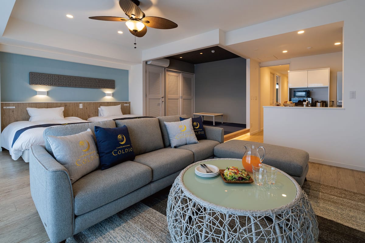 A modern hotel room with a bed, sofa, dining table, and a kitchenette.