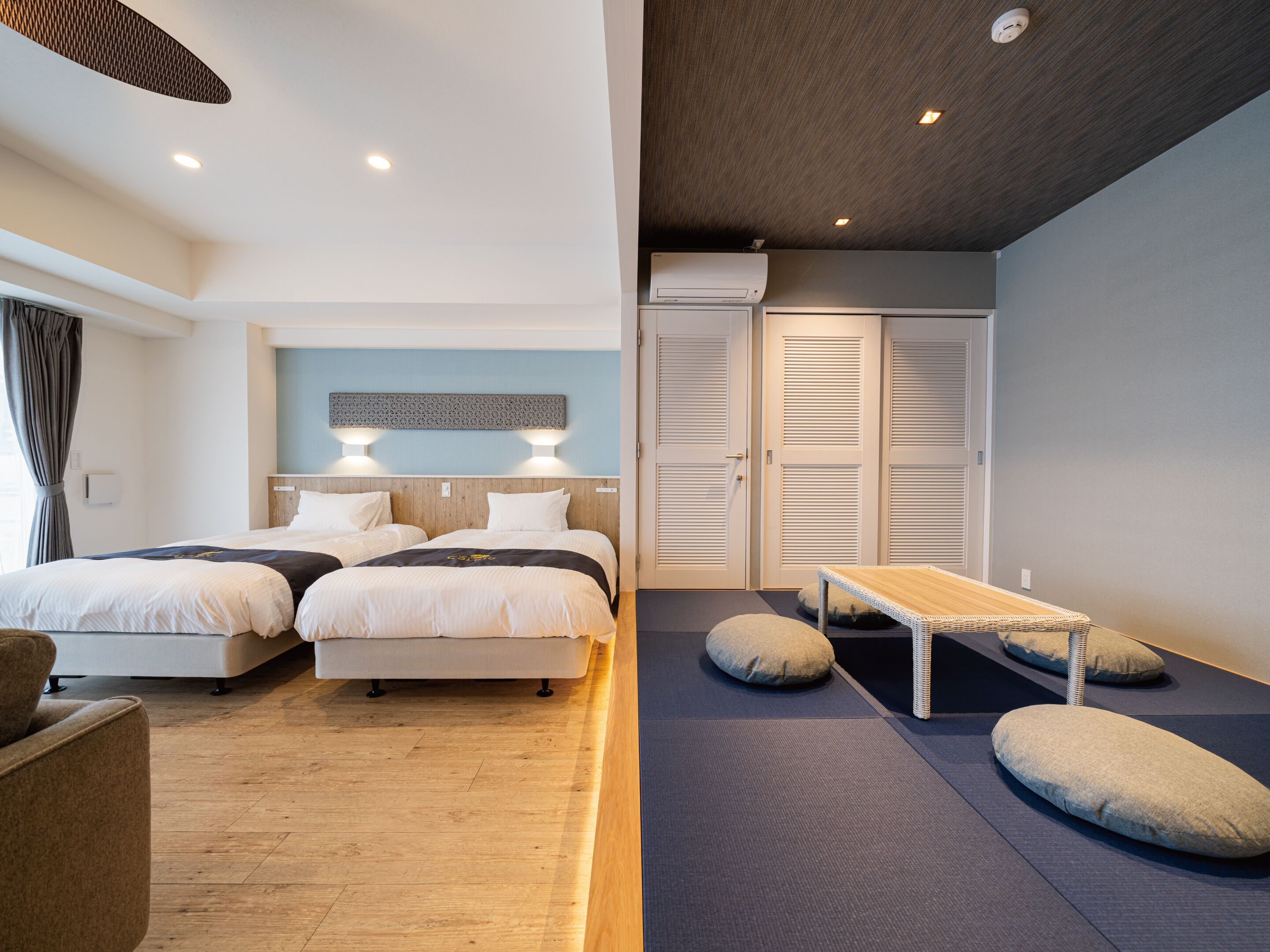 A hotel room with two beds, a small table, and a bench. There are ceiling lights and a wall-mounted air conditioner.