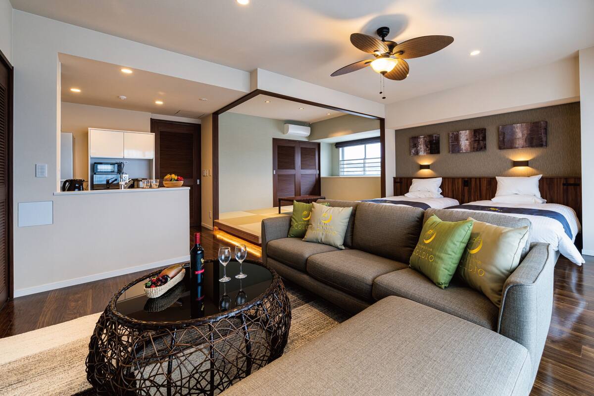 A modern hotel room with a sofa, a dining table, a ceiling fan, and a bed with a headboard.