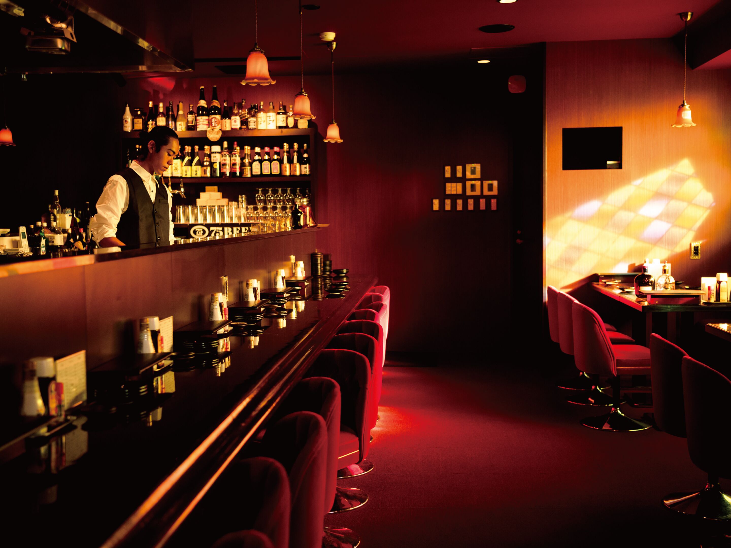 Un bar con un cantinero, estantes llenos de botellas de licor y taburetes rojos.