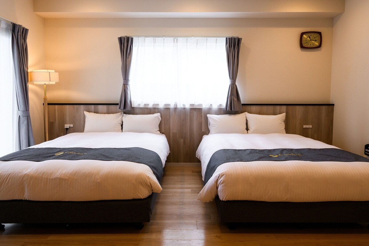 A hotel room with two beds, a wooden headboard, a window with curtains, a wall clock, and a bedside lamp.