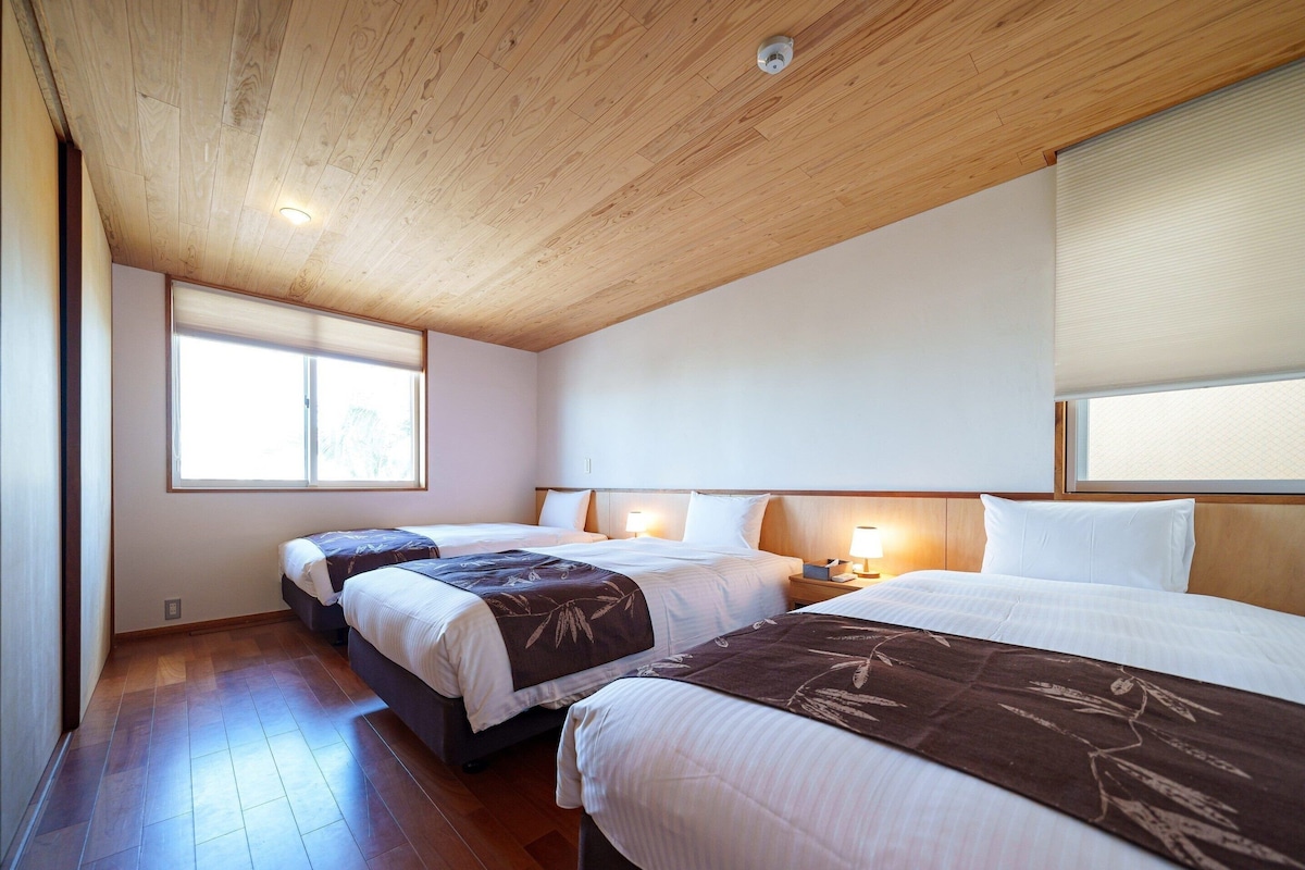 Habitación de hotel con dos camas, piso de madera, techo con vigas de madera y una ventana con persianas.