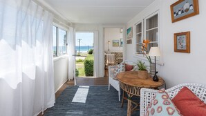 A cozy room with a wooden dining table, white wicker furniture, and a view of the sea through a door. - Original 1950's beach cottage (Mollymook)
