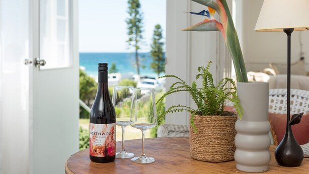 A table with a wine bottle, two glasses, a potted plant, a lamp, and a view of the ocean through a door. - Original 1950's beach cottage (Mollymook)