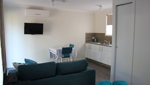 Een knus woongedeelte met een bank, een eettafel met stoelen, een keukenhoek en een aan de muur bevestigde televisie.