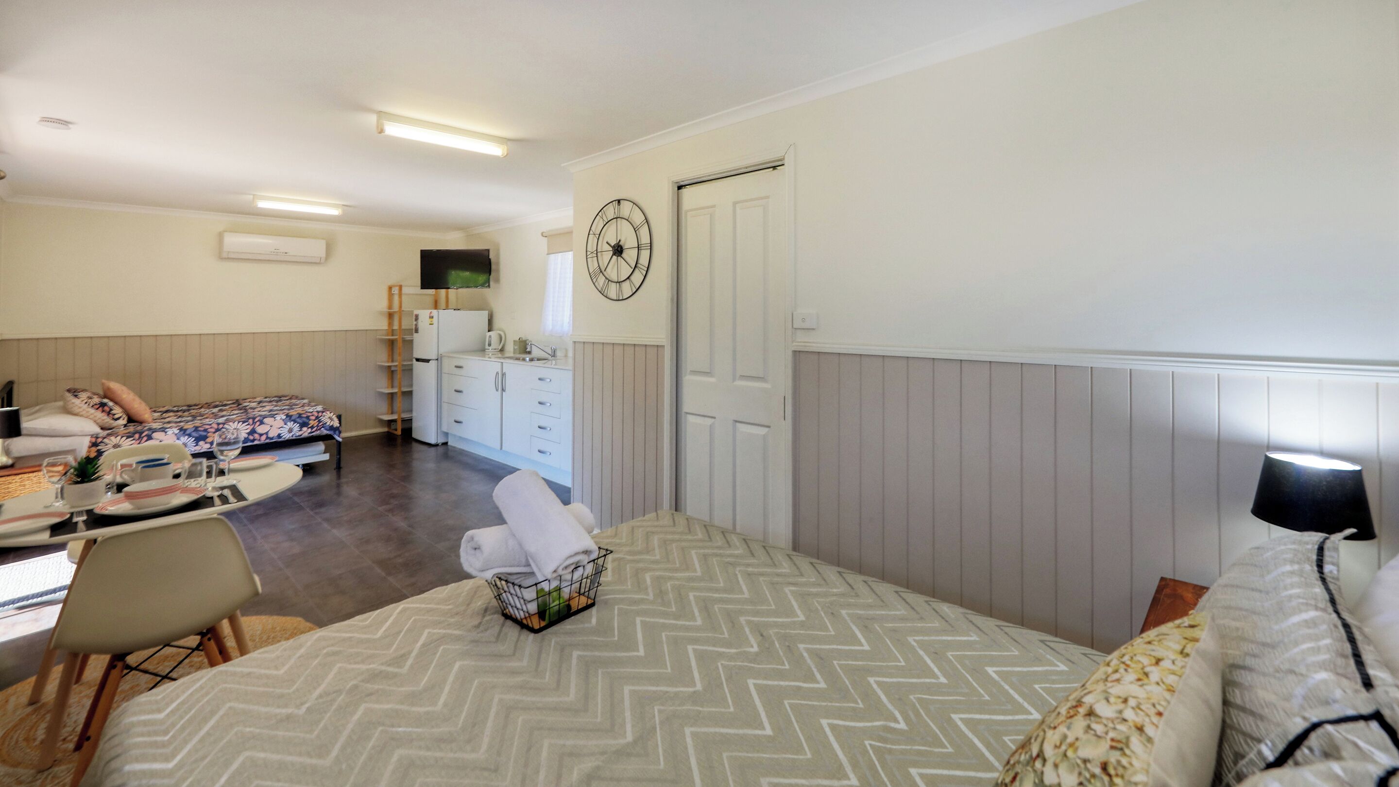 A hotel room with two beds, a kitchenette, a dining area, and a large wall clock.