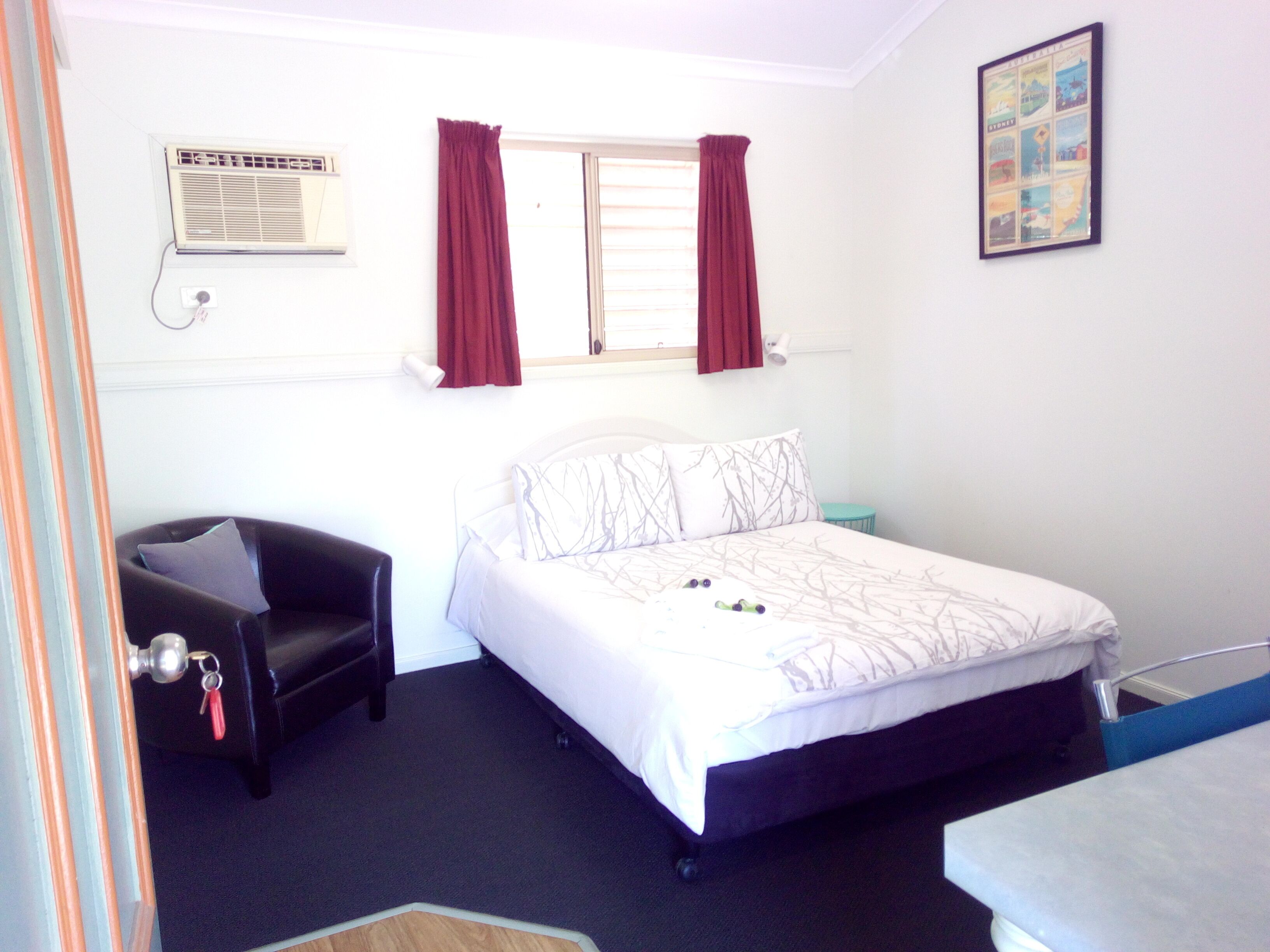 A bedroom with a bed, a chair, a small table, a window with curtains, and a wall-mounted air conditioning unit.