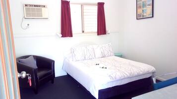 A bedroom with a bed, a chair, a small table, a window with curtains, and a wall-mounted air conditioning unit.