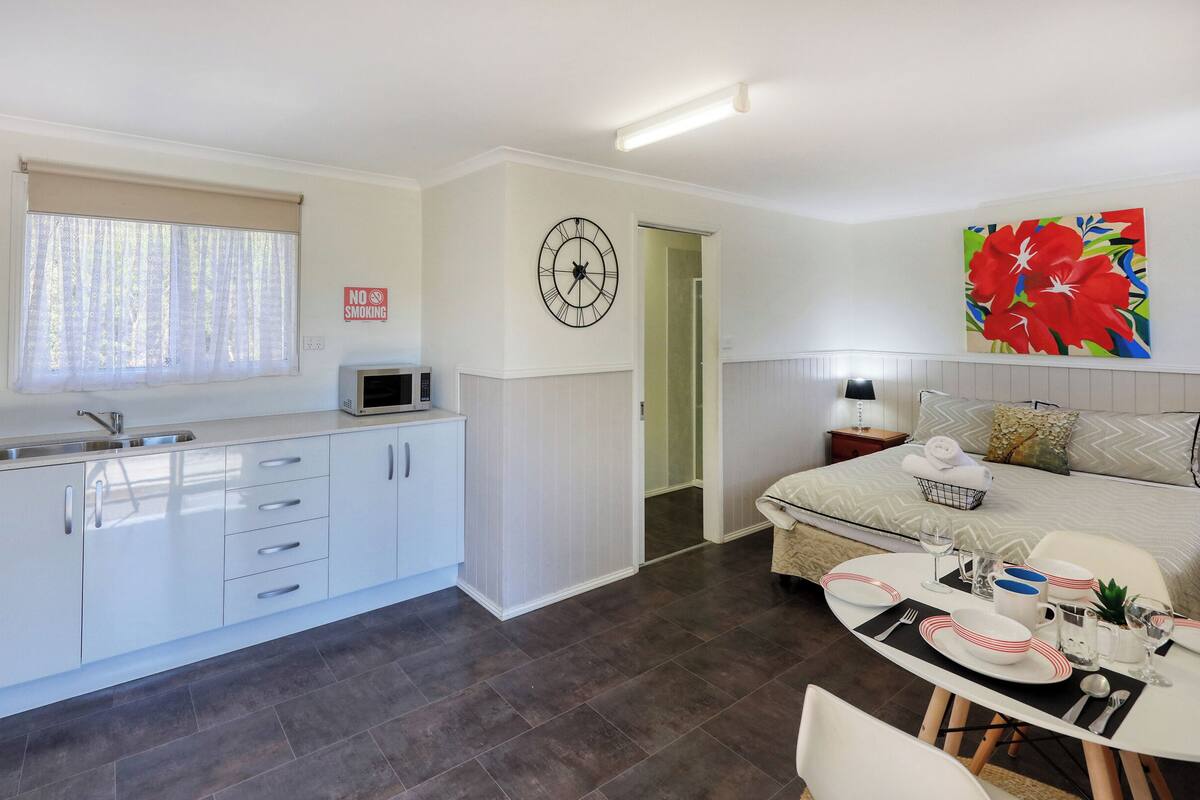 A modern kitchen with white cabinets, a microwave, and a sink. There is a dining area with a table set for two, a bed with pillows, and a colorful painting on the wall.