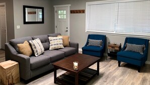 Un salon avec un canapé gris, des fauteuils bleus, une table basse et une fenêtre avec des stores.
