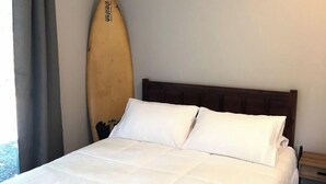 Un lit bien fait, avec une literie blanche, une tête de lit en bois et une planche de surf appuyée contre le mur.