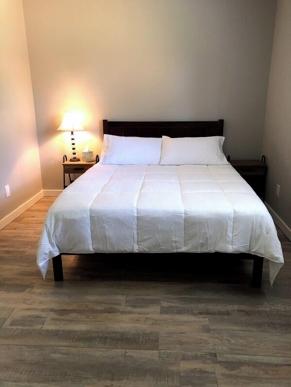 A neatly made bed with a white bedspread and two pillows, a bedside table with a lamp, and a wooden floor.