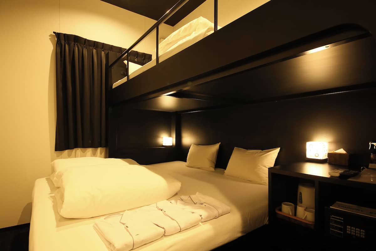A hotel room with a bunk bed, a bedside table with a lamp, and a shelf with cups and a remote control.