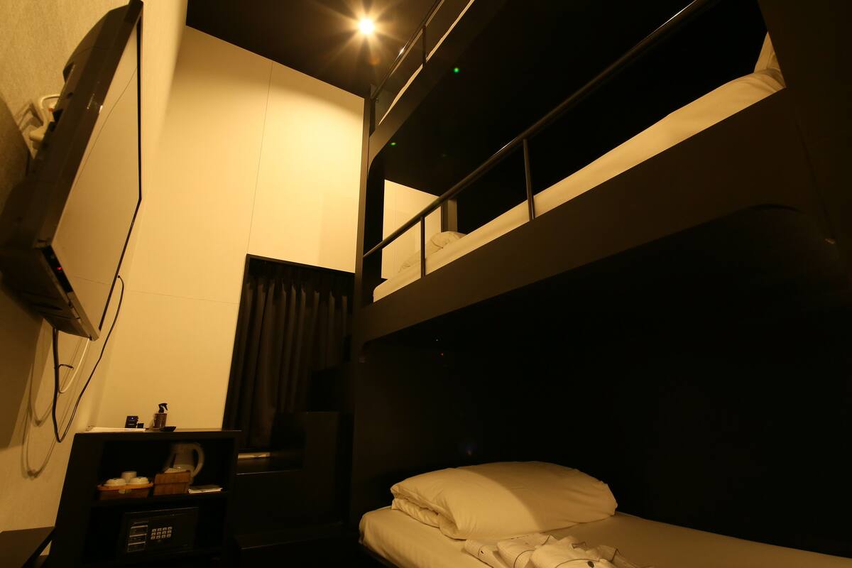 A compact hotel room with a bunk bed, a desk with a coffee maker, a TV, and a small shelf with toiletries.