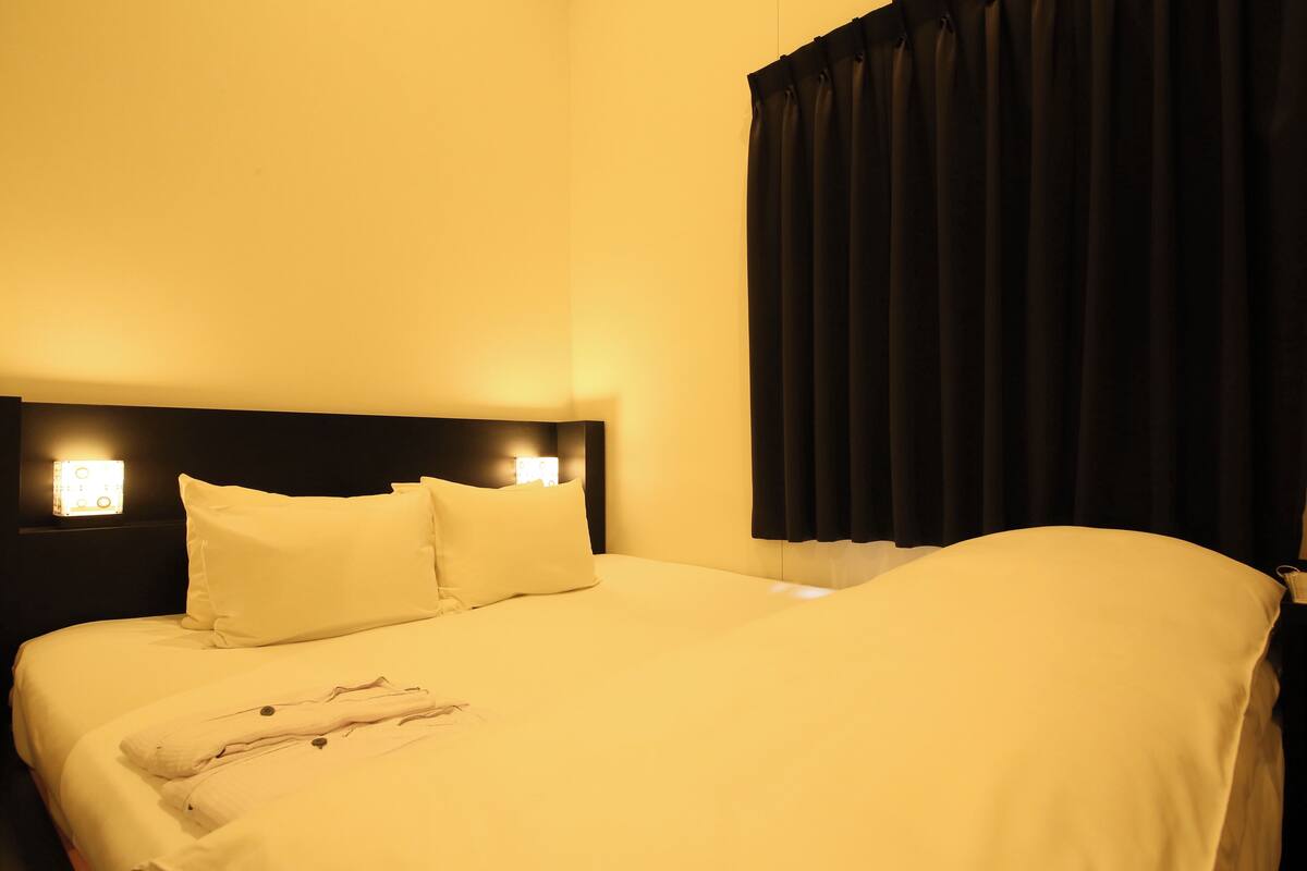 A neatly made bed with white linens and pillows, a headboard with wall-mounted lights, and a dark curtain on the window.