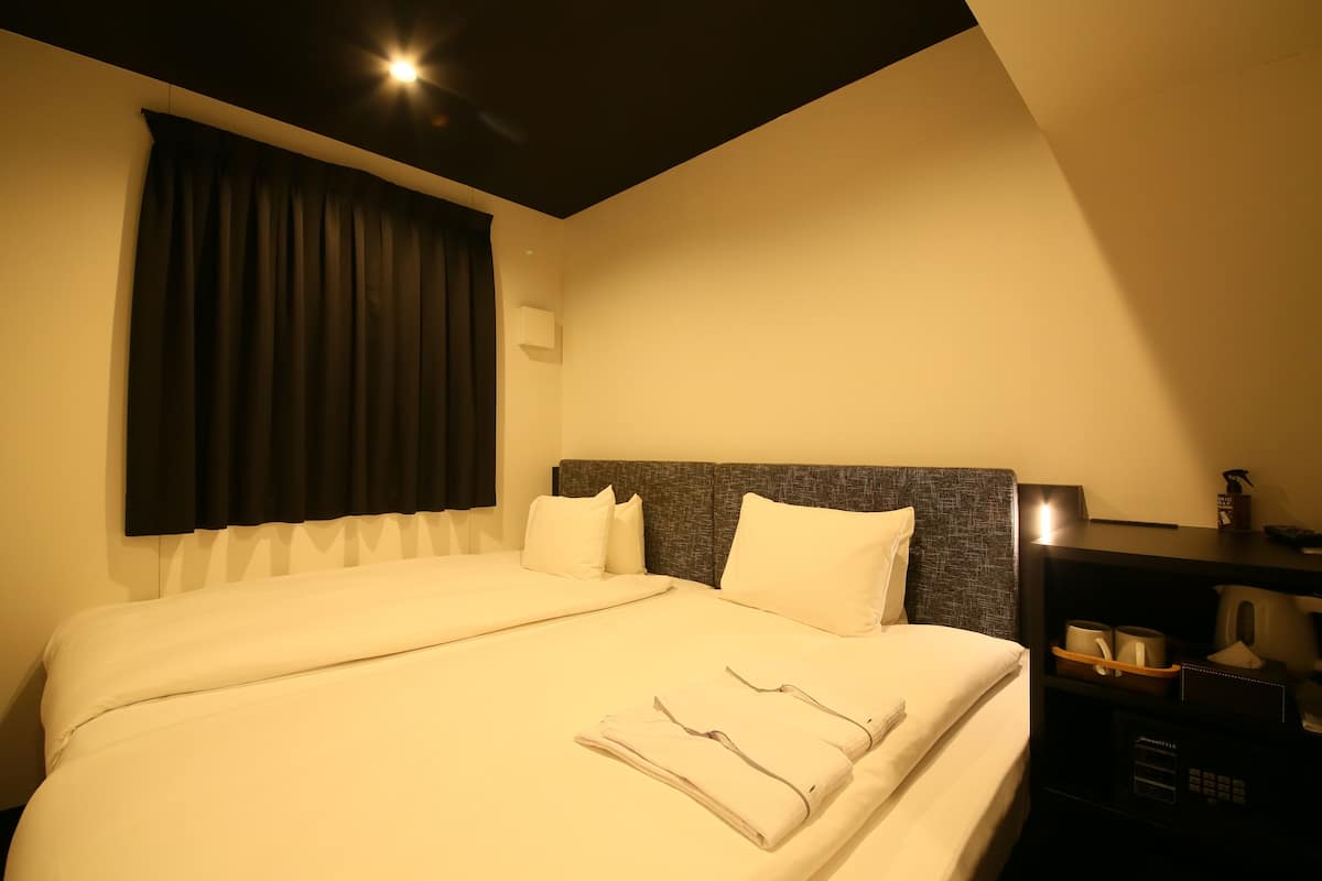 A hotel room with two beds, a dark headboard, a nightstand with a lamp, and a window with curtains.