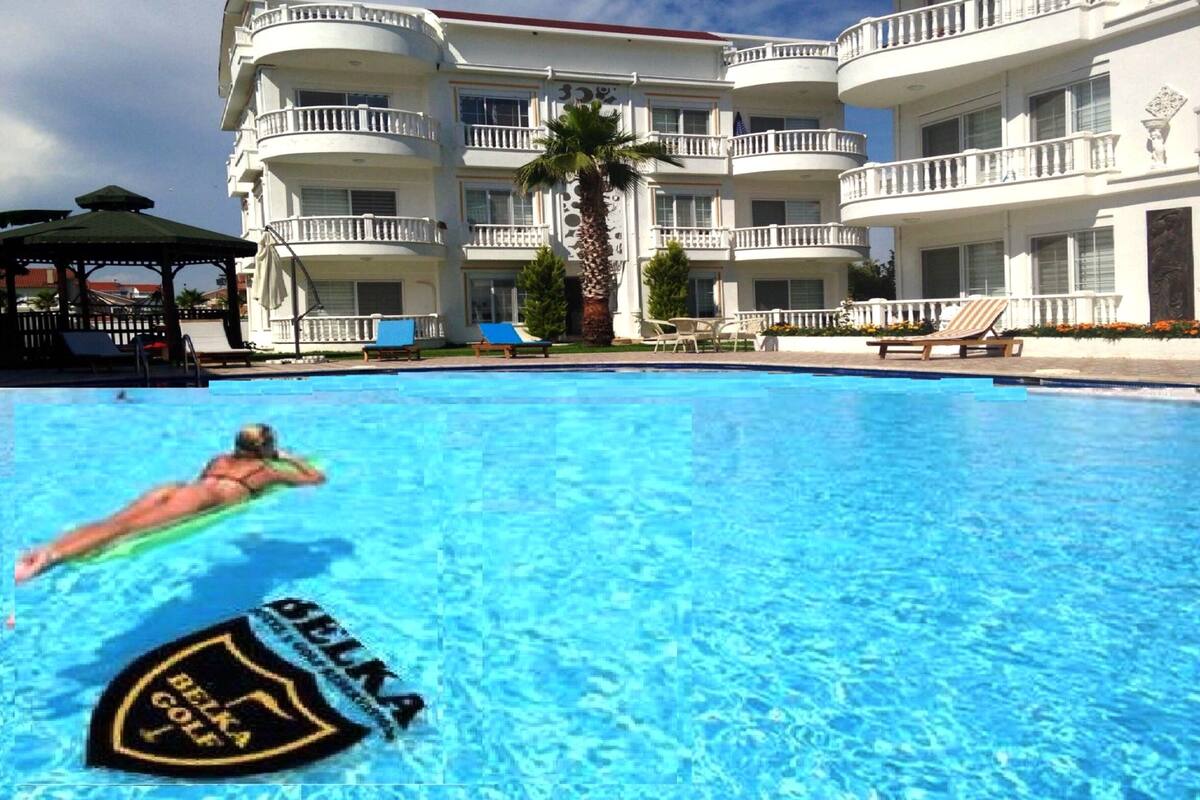 A swimming pool with a person swimming, surrounded by a multi-story building with balconies and a palm tree.