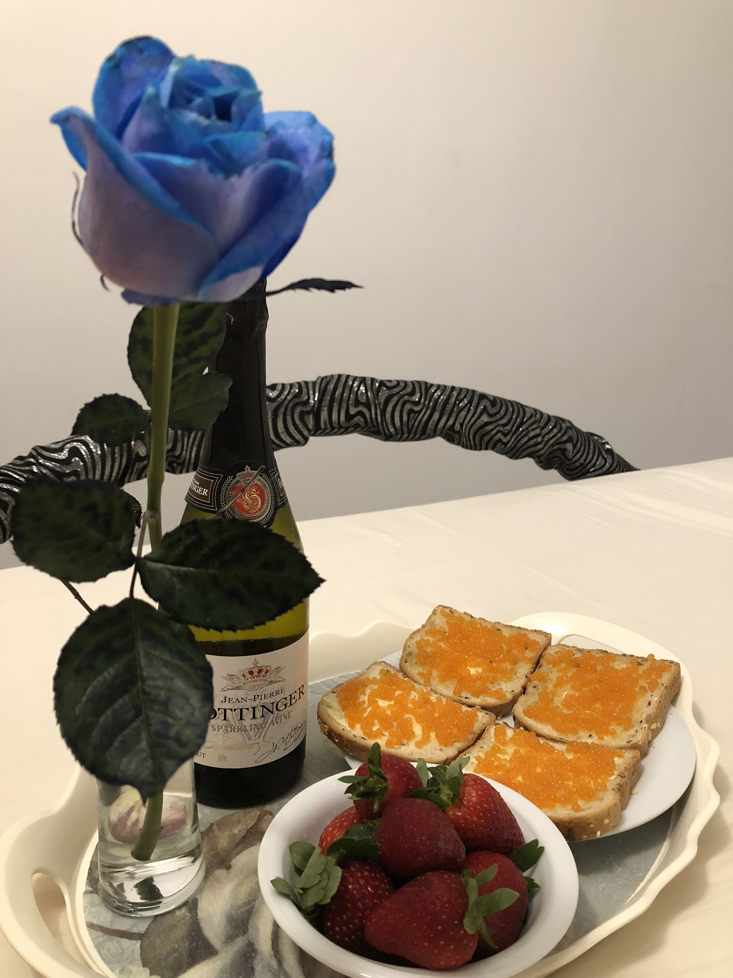 A table with a bottle of Jean-Pierre Pottinger sparkling wine, a blue rose, slices of bread with orange caviar, and a bowl of strawberries.