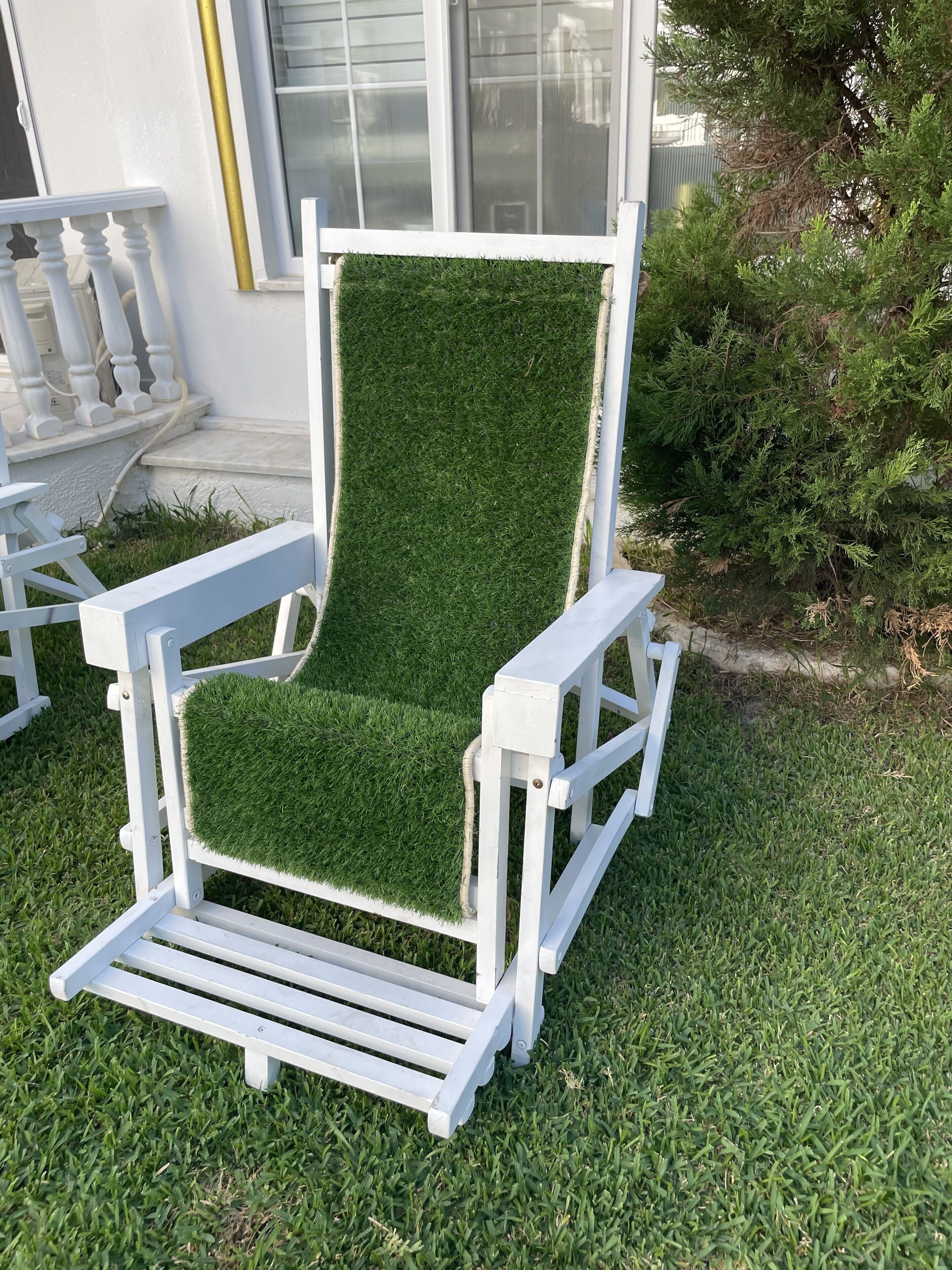 Une chaise d’extérieur blanche, dotée d’un siège et d’un dossier recouverts de gazon synthétique vert, est placée sur une pelouse, près d’un bâtiment blanc muni d’une fenêtre et d’une porte.