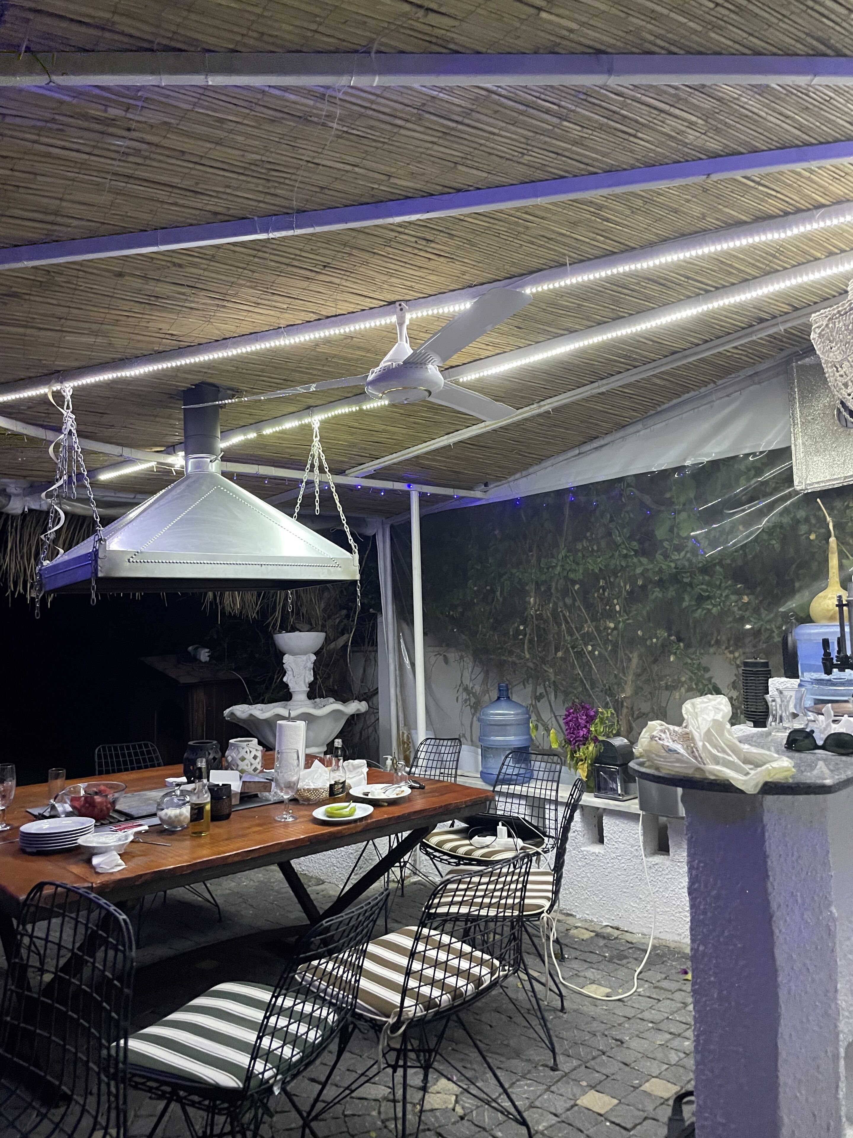 An outdoor dining area with a wooden table, metal chairs, hanging pendant lights, and a large water dispenser.