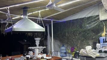 An outdoor dining area with a wooden table, metal chairs, hanging pendant lights, and a large water dispenser.