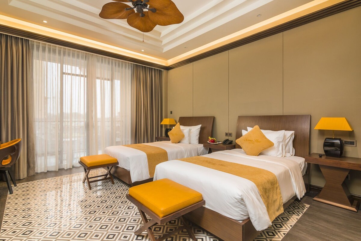 A hotel room with two beds, a desk, a chair, and a large window with curtains.