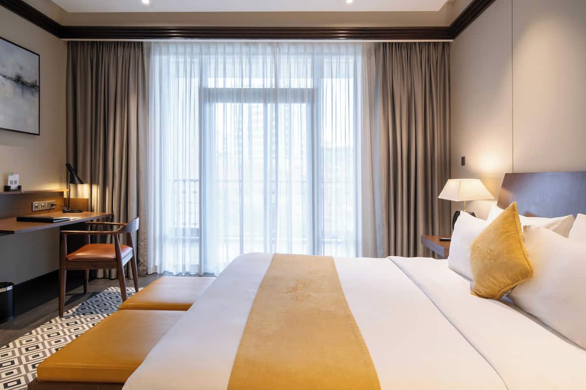 A hotel room with a large bed, a desk with a chair, a small table, and a lamp.