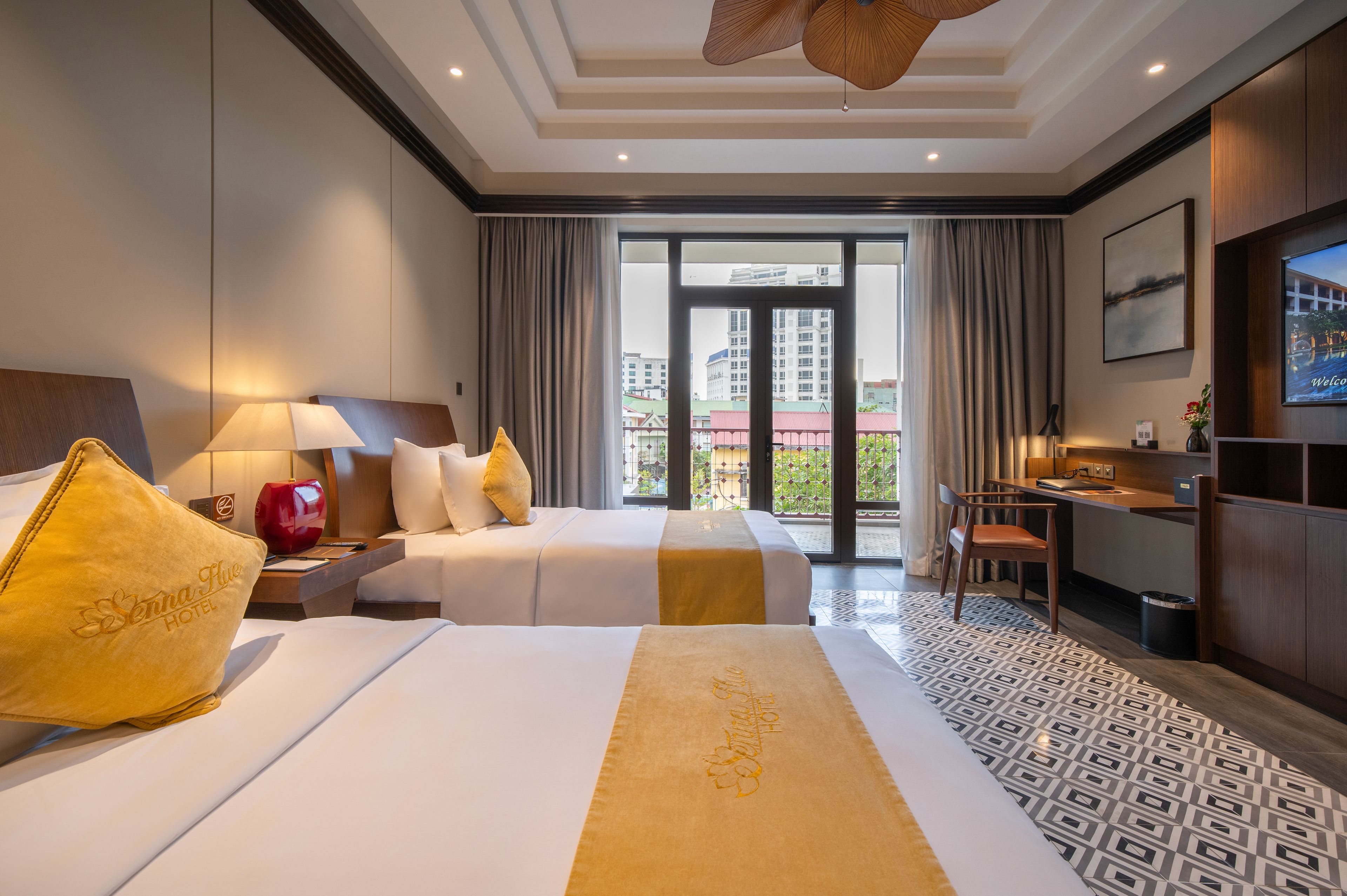 A hotel room with two beds, a desk, a TV, and a balcony with a city view.