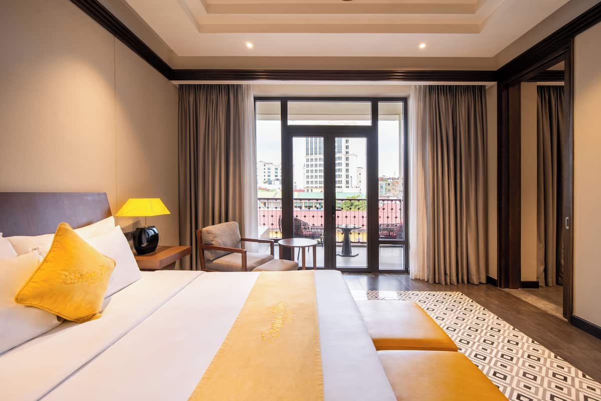 A hotel room with a bed, a nightstand, a lamp, a chair, a small table, and a view of the outside through a glass door.