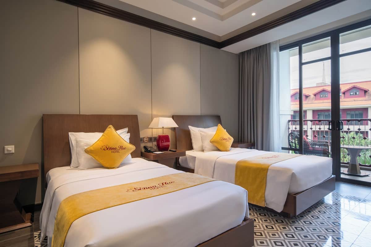 A hotel room with two beds, a nightstand with a lamp, and a view of buildings through large windows.