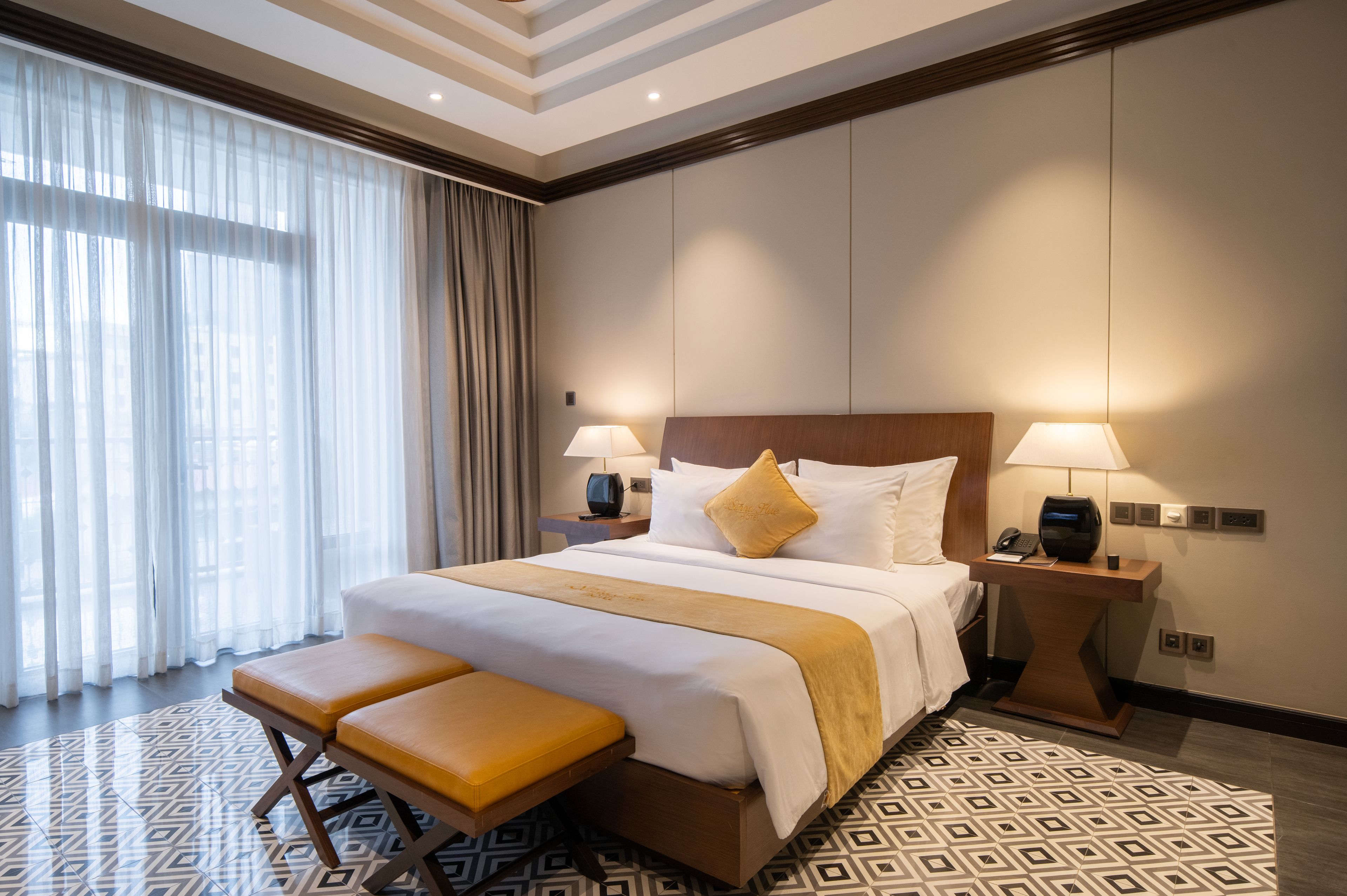 A hotel room with a large bed, two bedside tables, a bench, and a patterned floor.
