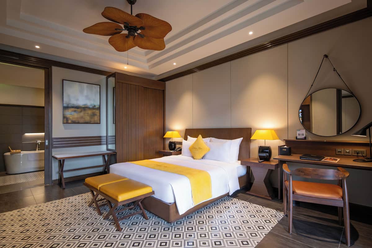 A hotel room with a large bed, bedside lamps, a desk, a chair, a bathroom, and a ceiling fan.