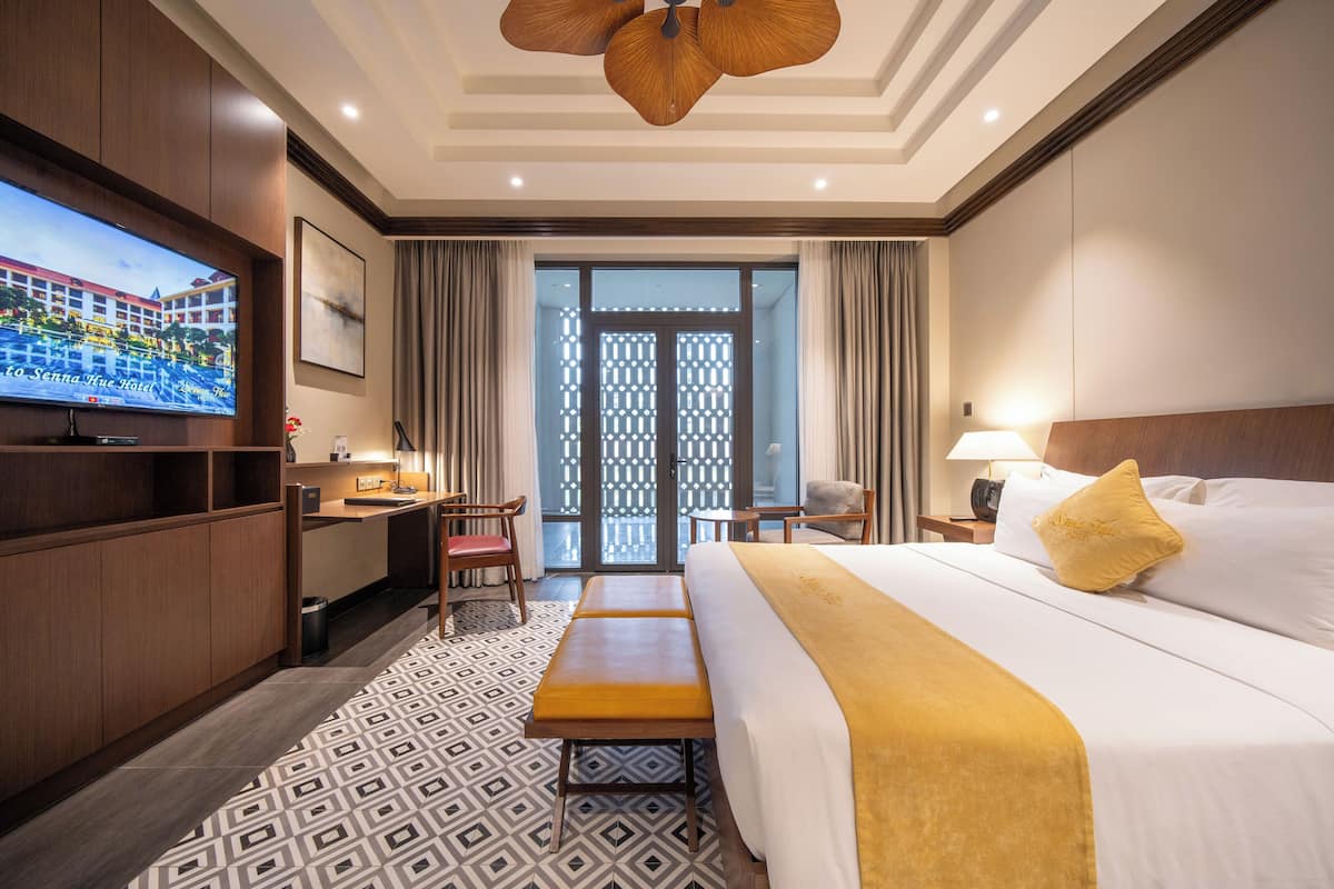 A hotel room with a large bed, a TV, a desk, and a bench.