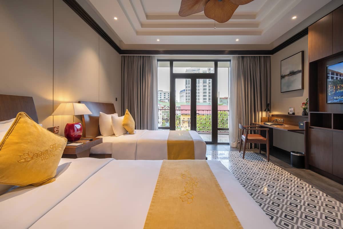 A hotel room with two beds, a desk, a TV, and a balcony with a city view.