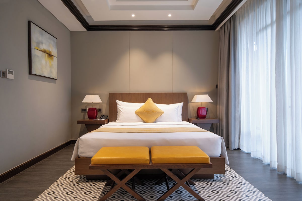 Ein Hotelzimmer mit einem großen Bett, zwei Nachttischlampen, einem Nachttisch, einem gemusterten Teppich und einem großen Fenster mit Vorhängen.