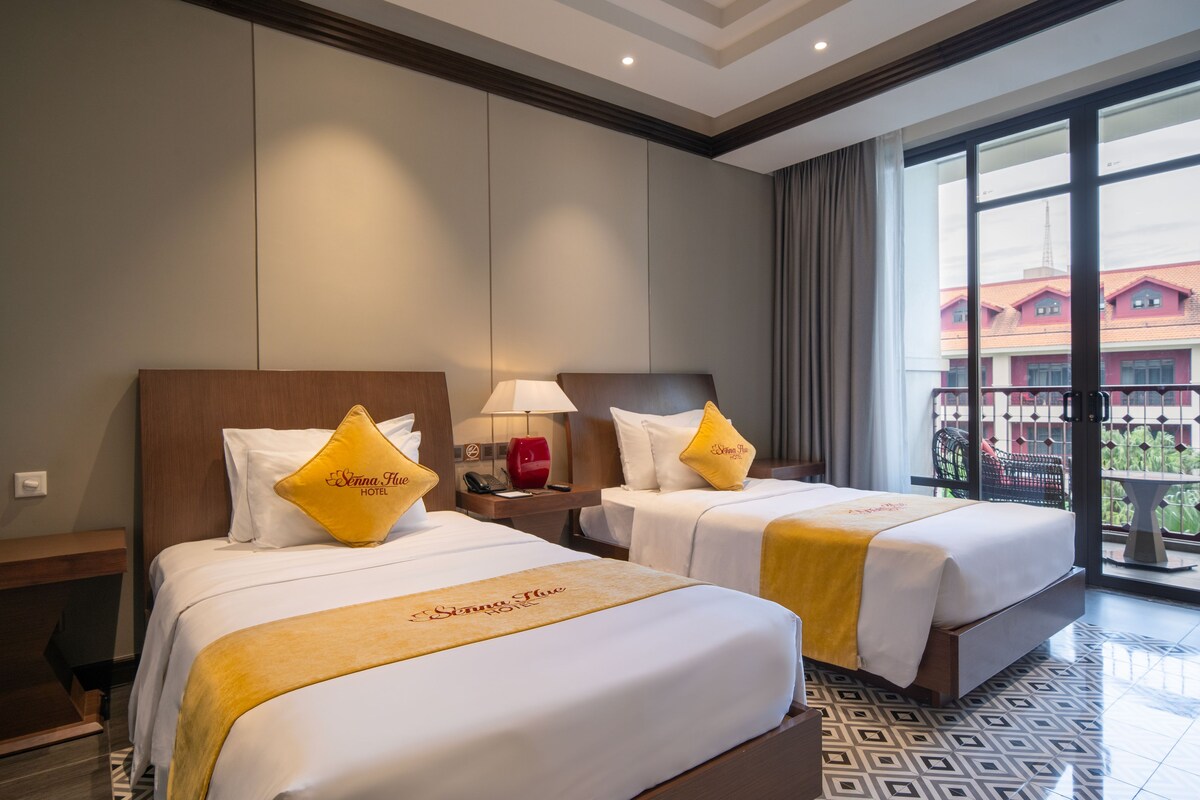 A hotel room with two beds, a nightstand with a lamp, and a view of a building through large windows.