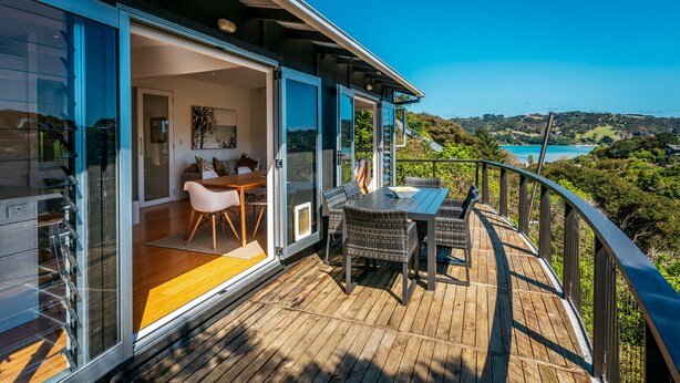 A modern house with a wooden deck, a dining area with wicker furniture, and a large glass door leading to a living room.