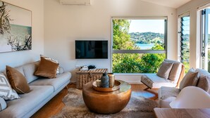 A modern living room with a sofa, armchairs, a coffee table, and a TV. There is a large window with a view of greenery and a body of water.