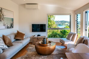 Uma sala de estar moderna com um sofá, poltronas, uma mesa de centro e uma TV. Há uma janela grande com vista para áreas verdes e um corpo d'água.