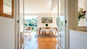 A modern interior with a wooden floor, a dining area with white chairs, and a view of greenery through large glass doors.