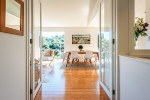 Um interior moderno com piso de madeira, área de refeições com cadeiras brancas e vista para o verde através de grandes portas de vidro.