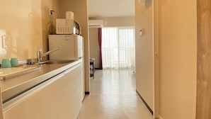 A narrow, well-lit hallway with a kitchen area on the left, featuring a sink, a microwave, and a refrigerator. The hallway leads to a room with a large window and a door on the right. - Popular Makiminato (Urasoe)
