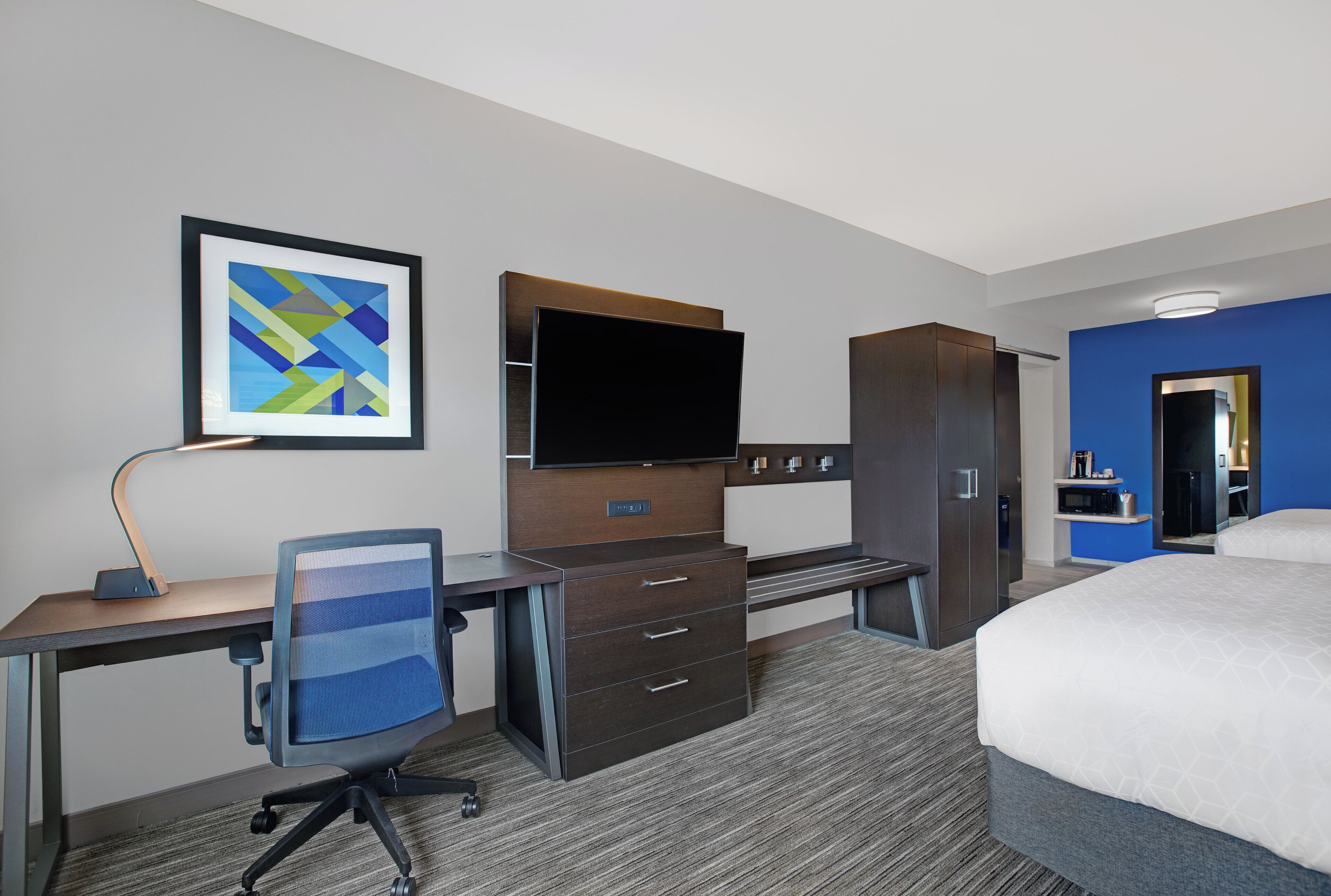 Une chambre d’hôtel comprenant un lit, un bureau avec une chaise, un téléviseur à écran plat, une armoire, un tableau encadré au mur et une salle de bain visible à travers une porte ouverte.