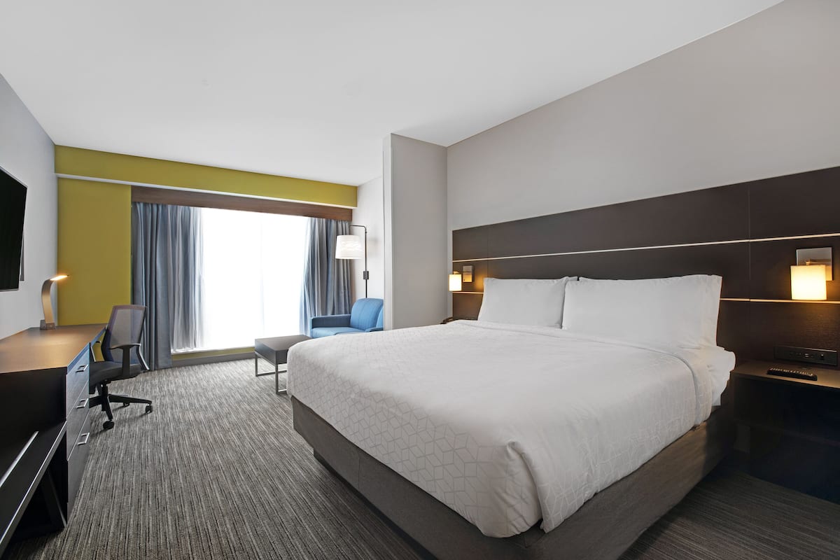 A hotel room with a large bed, a desk with a chair, a TV, a lamp, and a window with curtains.
