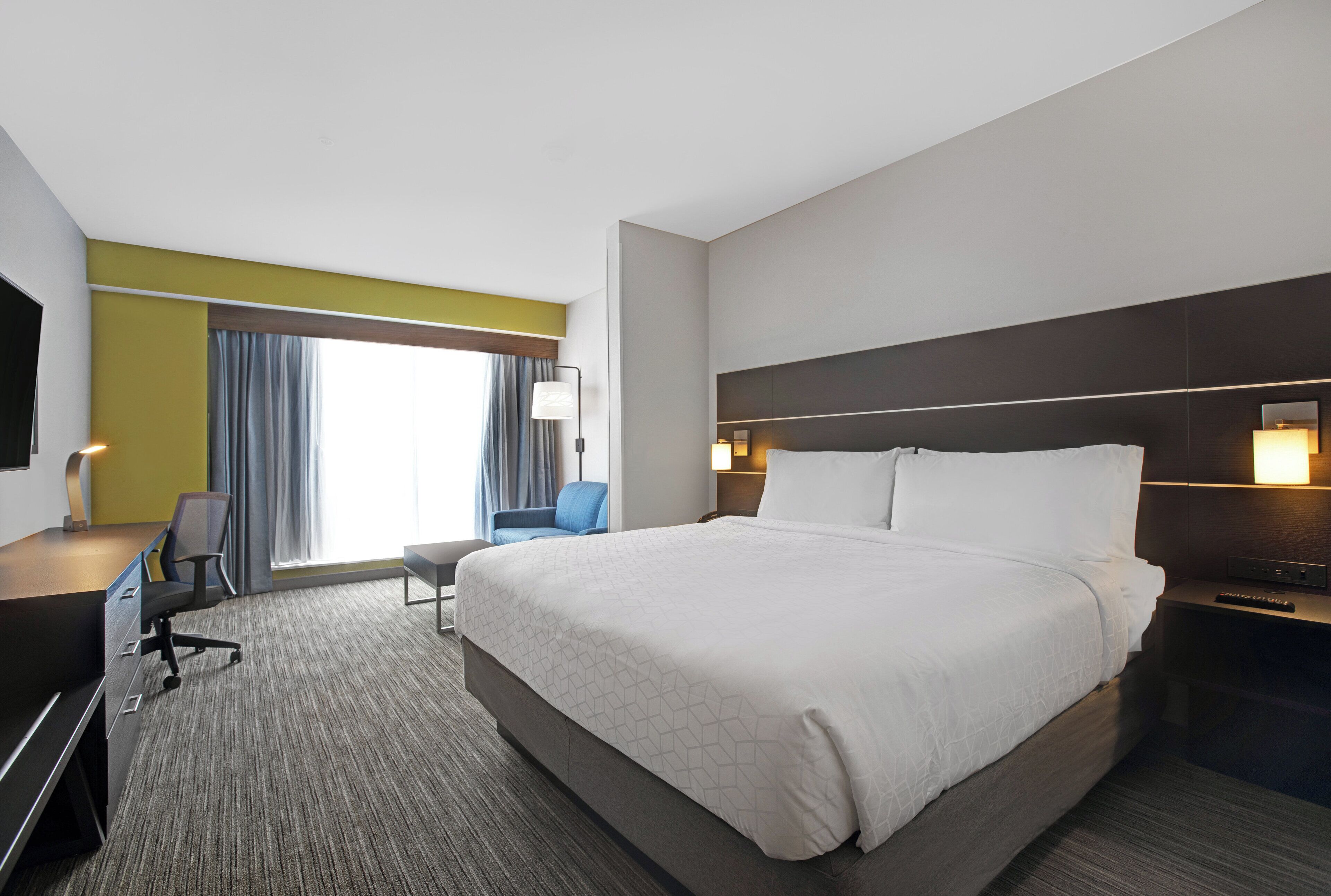 A hotel room with a large bed, a desk, a chair, a TV, and a window with curtains.