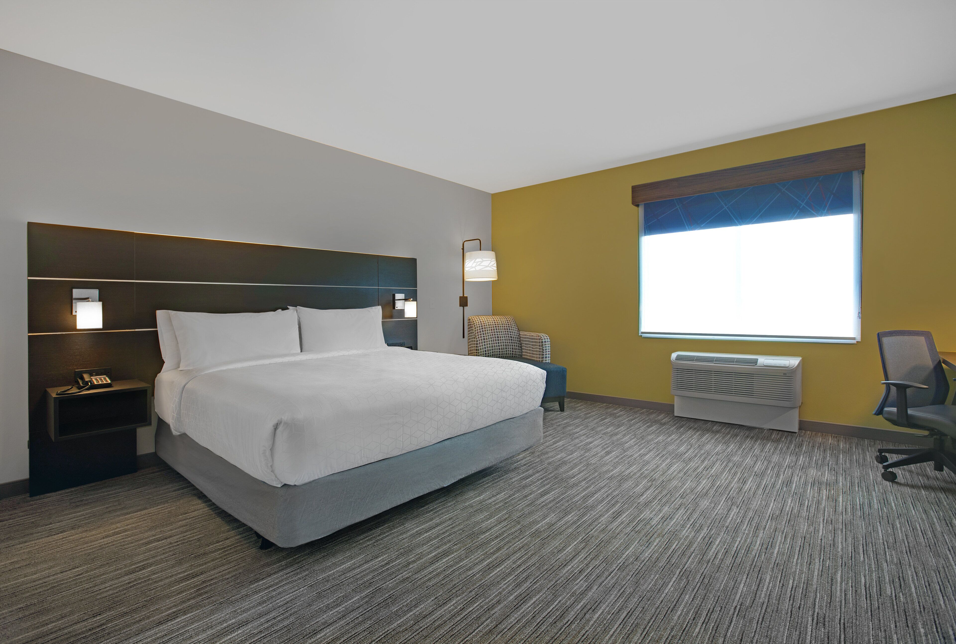 Une chambre d’hôtel avec un lit, une table de chevet, une lampe, une chaise et une fenêtre avec des stores.